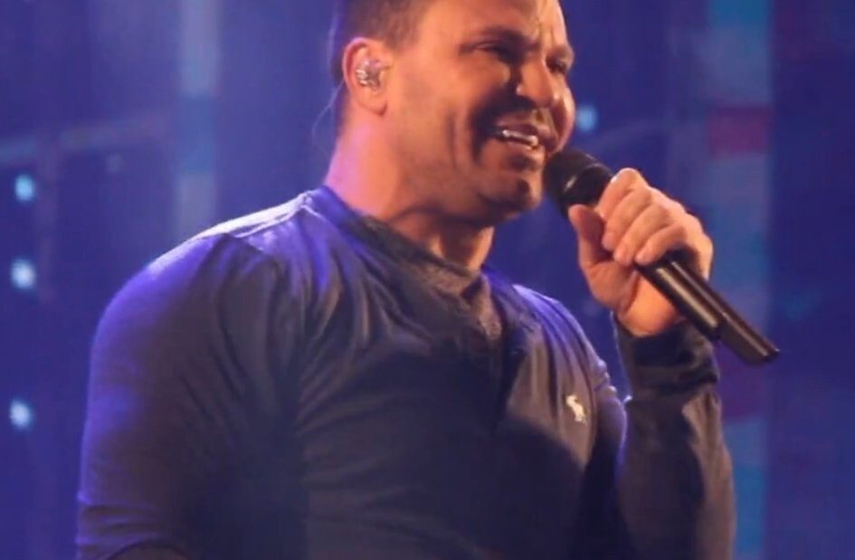 O cantor Eduardo Costa, de camiseta escura, sorri e canta ao microfone em um palco iluminado por luzes azuis