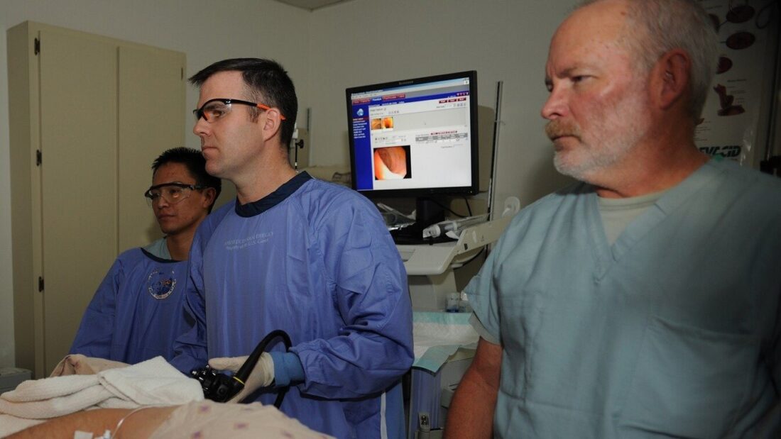 Médico de avental azul e óculos de proteção segura um endoscópio flexível enquanto observa um monitor com imagens do exame, ao lado de outros dois profissionais de saúde
