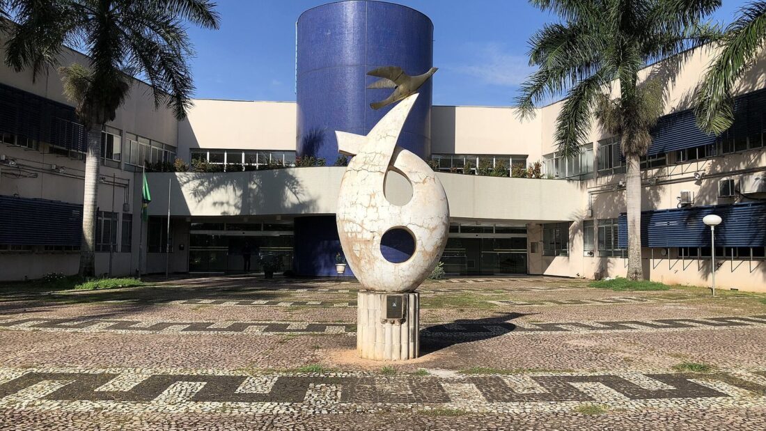 Prédio da Reitoria da Universidade Federal de Goiás, com torre cilíndrica azul, escultura branca à frente e palmeiras