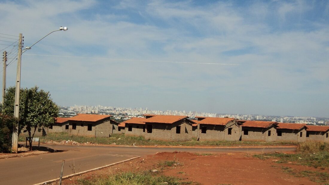 Rua de terra e asfalto em frente a um conjunto de casas térreas com telhado de cerâmica, com prédios de Goiânia ao fundo sob céu azul