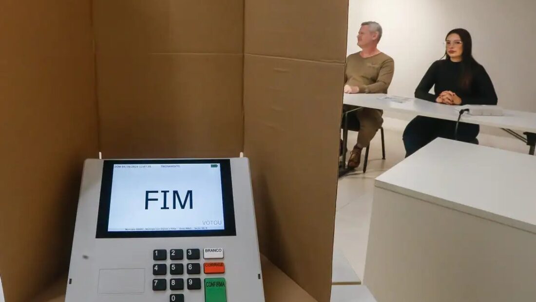 Urna eletrônica com a palavra FIM na tela, dentro de cabine de votação de papelão, com mesários sentados ao fundo