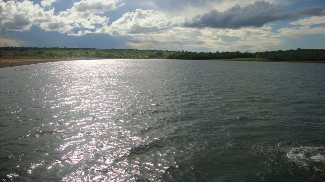 Lago de São Simão, com a água refletindo o sol, margem com vegetação ao fundo e céu com nuvens