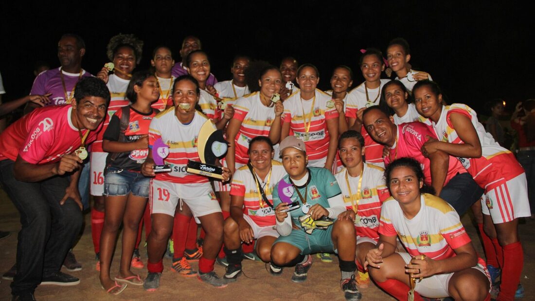 Jogadoras de um time de futebol feminino, de uniforme vermelho e branco, comemoram com medalhas e um troféu à noite, à beira do campo