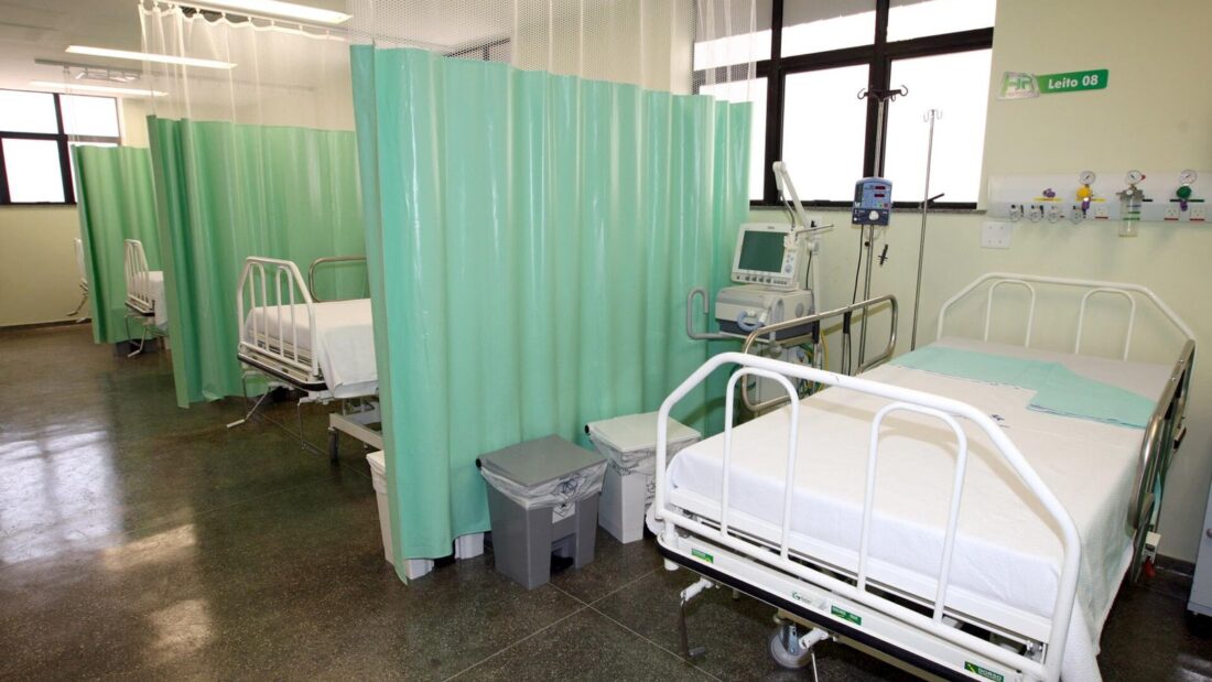 Leitos de UTI separados por cortinas verdes, com monitores e equipamentos junto às camas