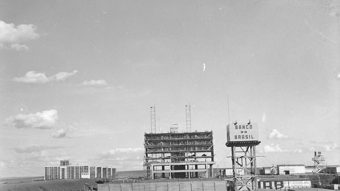 Fotografia antiga em preto e branco de um prédio em construção, com a estrutura de concreto exposta, andaimes e uma torre ao lado, em terreno aberto