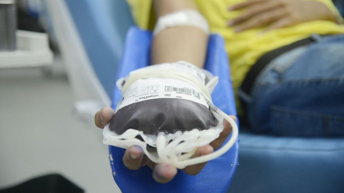 Mão de doador segurando bolsa de sangue recém-coletada, apoiada em cadeira de doação