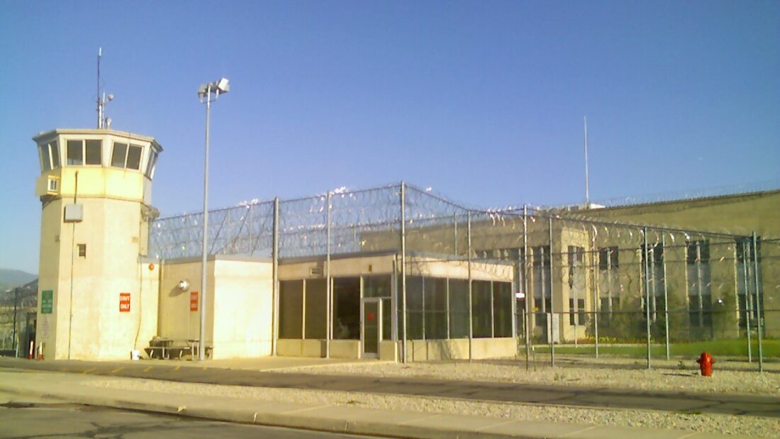 Entrada de visitantes de um presídio, com prédio baixo cor de areia, torre de vigilância e cercas com arame farpado, sob céu azul