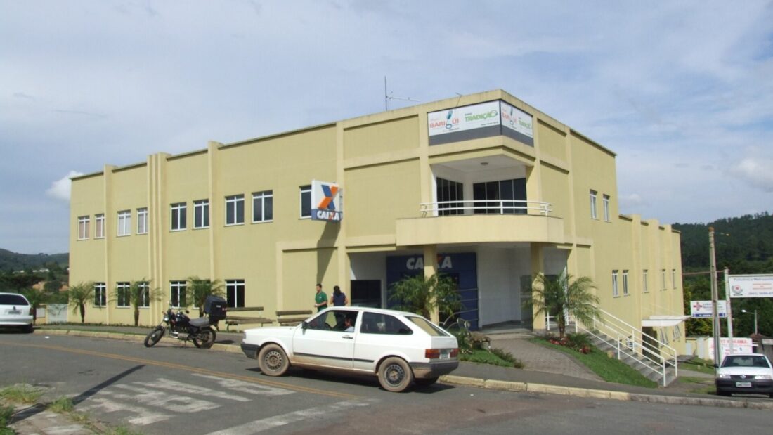 Prédio de dois andares pintado de amarelo-claro com o logotipo da Caixa Econômica Federal na fachada, na esquina de uma rua, com carros estacionados