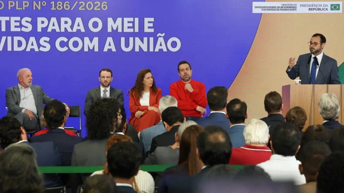 Presidente Lula e ministros sentados em um palco diante de um painel azul com o texto do PLP 186/2026, que trata do MEI e de dívidas com a União, enquanto um homem discursa no púlpito para uma plateia
