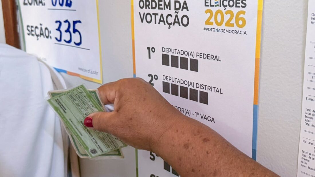 Mão de eleitora segura o título diante de cartazes com o número da seção e a ordem de votação das Eleições 2026