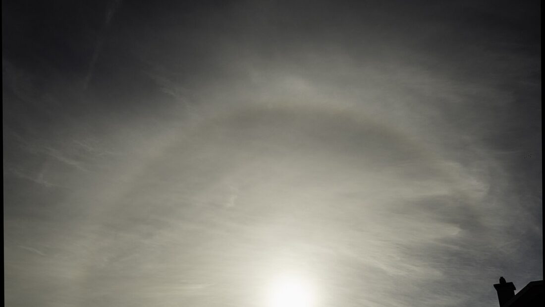 Céu coberto por nuvens finas e altas com um grande anel de luz em volta do Sol, acima da silhueta escura de telhados