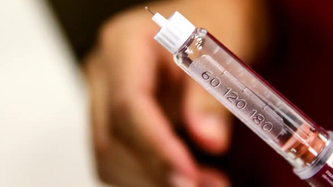 Mão segurando uma caneta aplicadora transparente com escala de doses marcada em números, em fundo desfocado