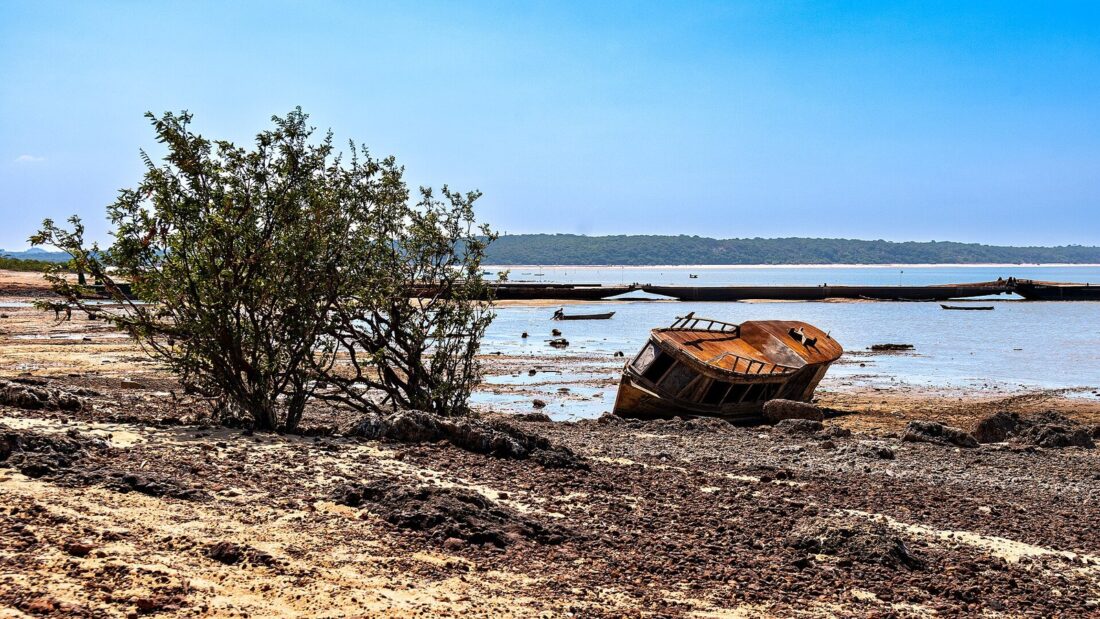 Casco de barco enferrujado encalhado na margem seca de um rio largo, ao lado de um arbusto