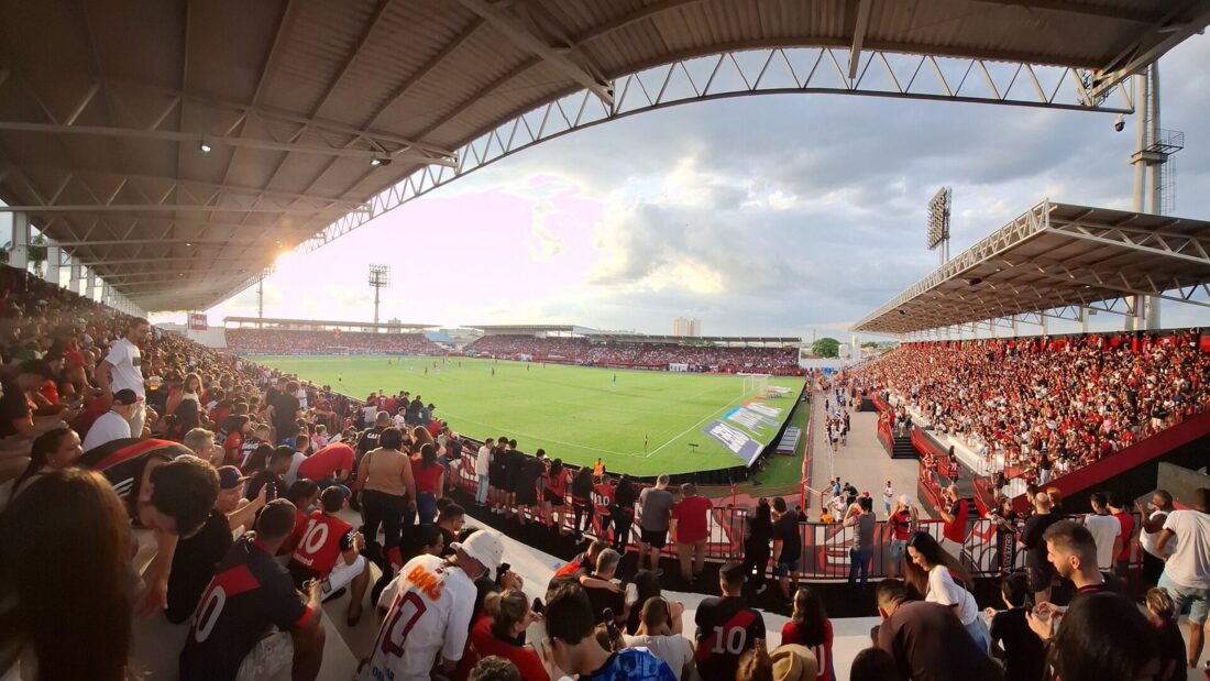 Arquibancada cheia de torcedores com camisas vermelhas e pretas no Estádio Antônio Accioly, com o gramado e a cobertura metálica ao fundo