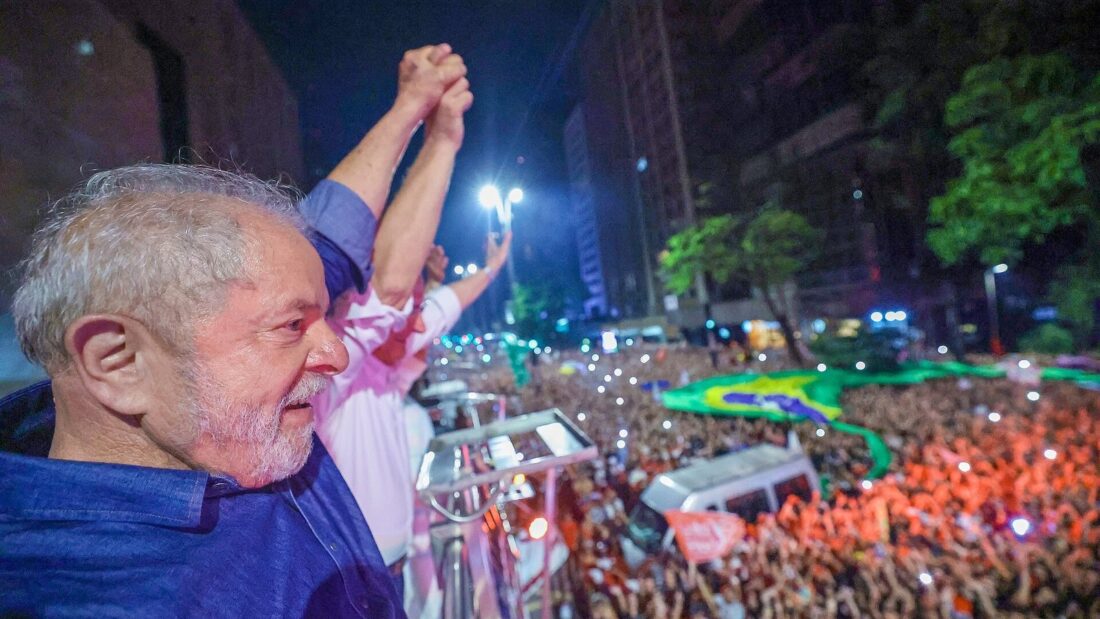 Lula, de camisa azul, de perfil diante de uma multidão à noite em uma avenida iluminada, com uma bandeira do Brasil estendida sobre o público