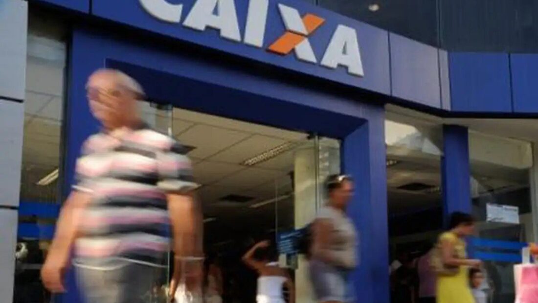 Pessoas passam em frente à entrada de uma agência da Caixa Econômica Federal, com o letreiro azul e laranja do banco no alto