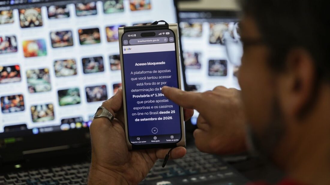 Pessoa segura celular com aviso de acesso bloqueado a plataforma de apostas, diante de uma tela de computador com jogos