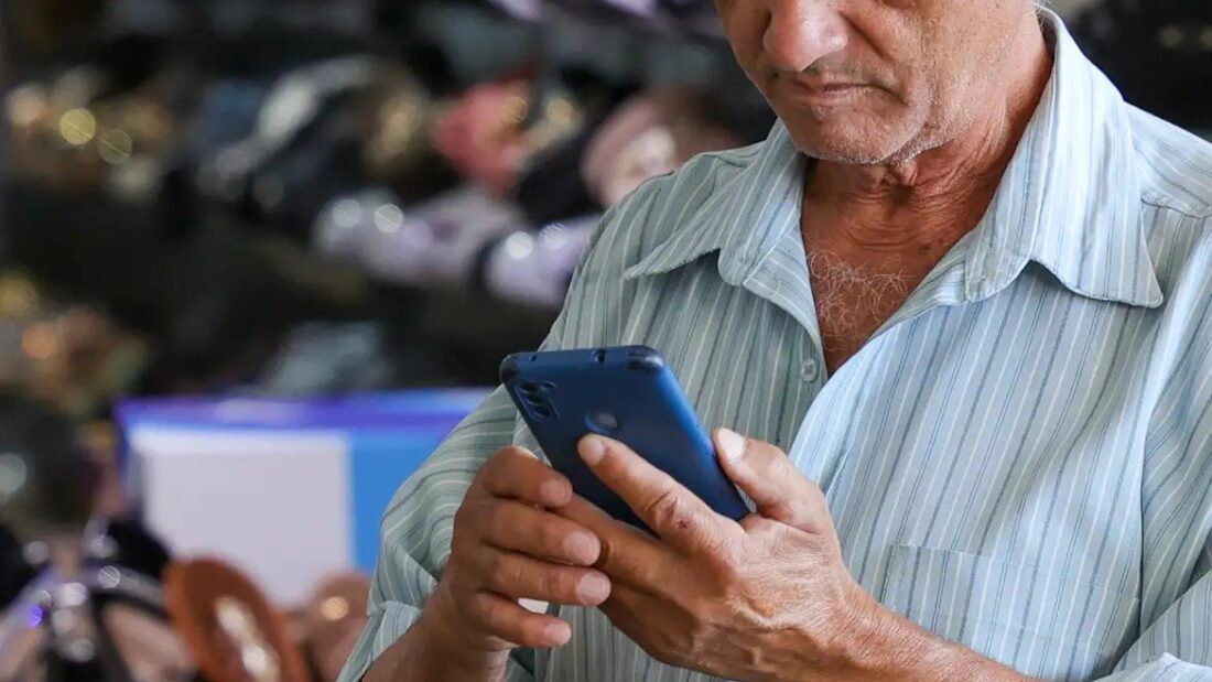 Homem idoso de camisa listrada olha para a tela do celular que segura com as duas mãos