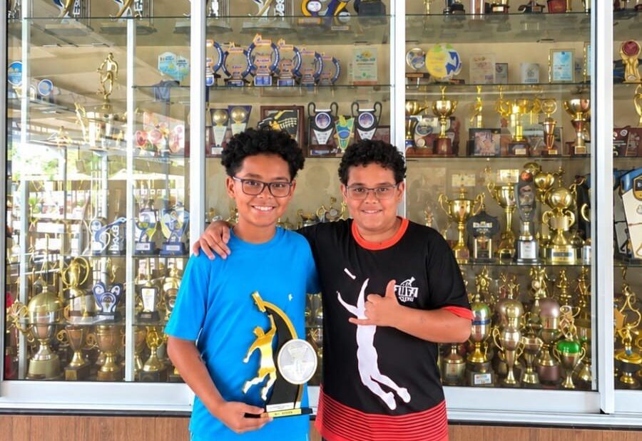 Dois meninos abraçados, um de camiseta azul segurando um troféu e outro de camiseta preta, diante de uma galeria de troféus