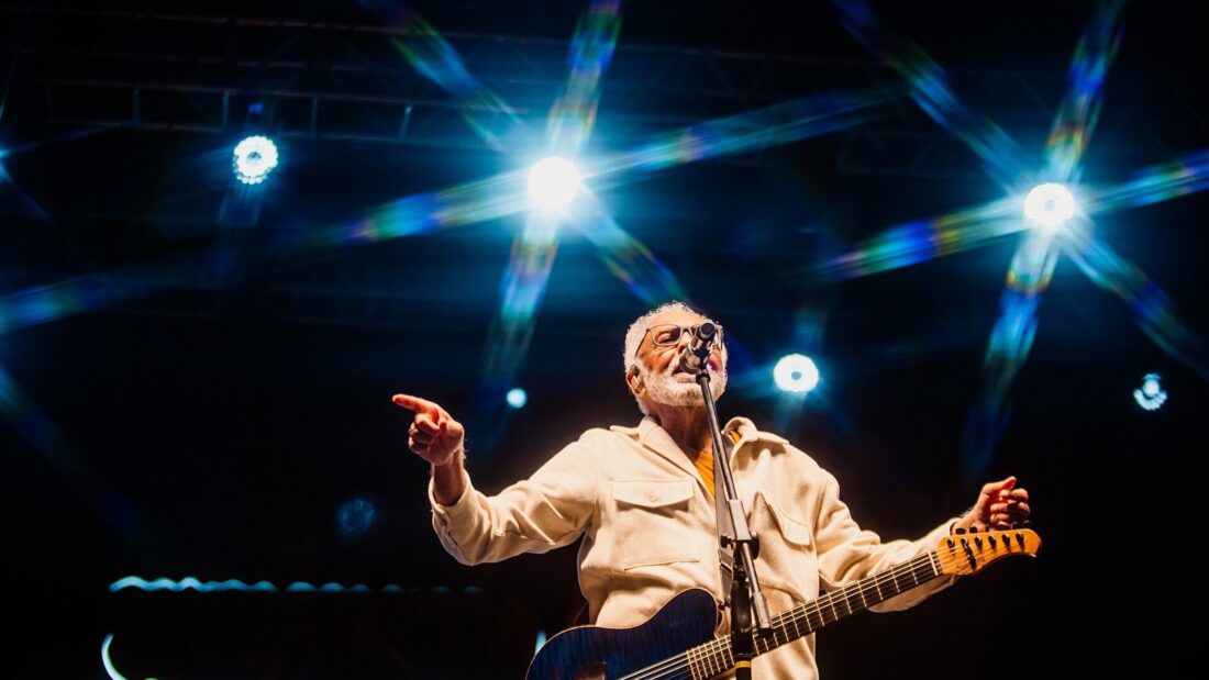 Gilberto Gil canta e toca violão em um palco iluminado por refletores azuis