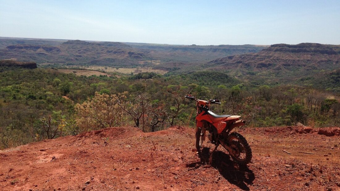 Vista do alto de um morro de terra vermelha, com uma motocicleta parada, sobre um vale de cerrado e serras ao fundo, na região de Caiapônia