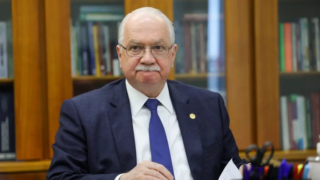 Edson Fachin, de óculos, cabelos e bigode brancos, terno escuro e gravata azul, sentado a uma mesa em frente a uma estante de livros