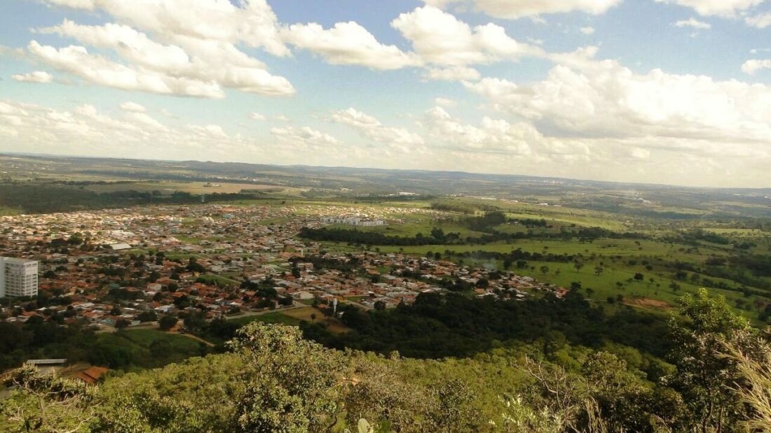 Vista do alto de um morro com vegetação de cerrado em primeiro plano e a área urbana de Senador Canedo espalhada ao fundo sob céu com nuvens