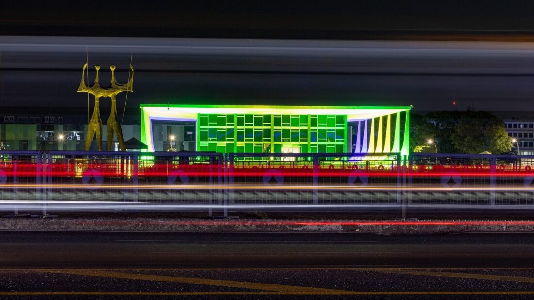 Palácio do Supremo Tribunal Federal iluminado à noite em Brasília, com a escultura dos Guerreiros à esquerda e rastros de luz de carros em primeiro plano
