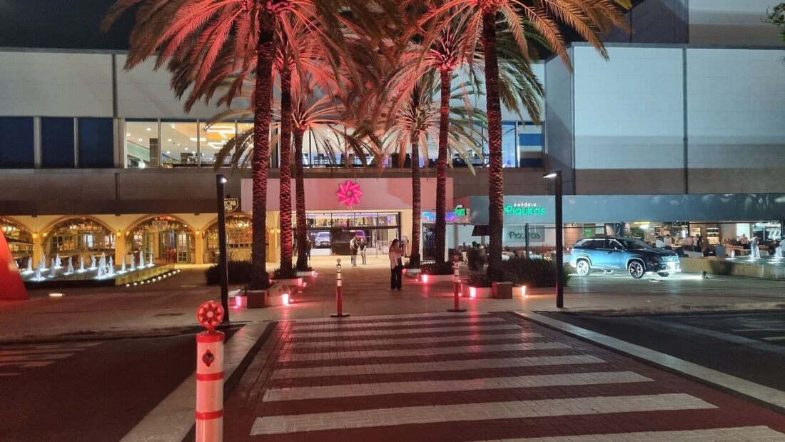 Entrada de um shopping center à noite, com palmeiras iluminadas e faixa de pedestres em primeiro plano