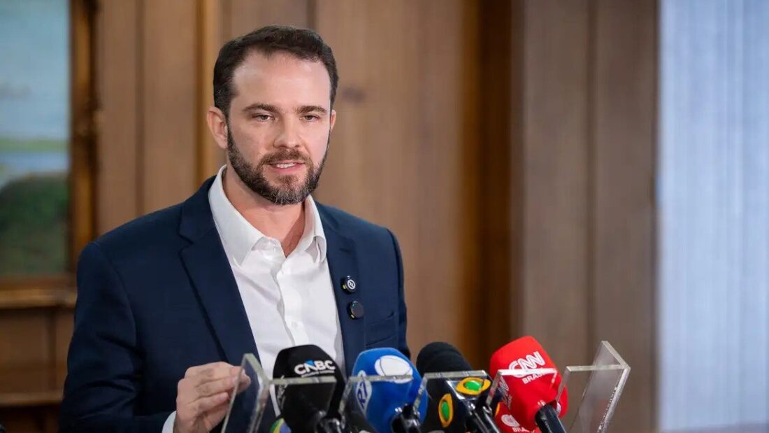 O ministro da Fazenda, Dario Durigan, de terno escuro e camisa branca, fala diante de microfones de emissoras de TV