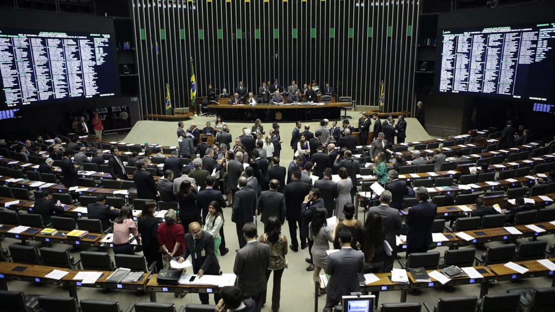 Plenário da Câmara dos Deputados visto do alto, com parlamentares em pé entre as bancadas e painéis de votação acesos