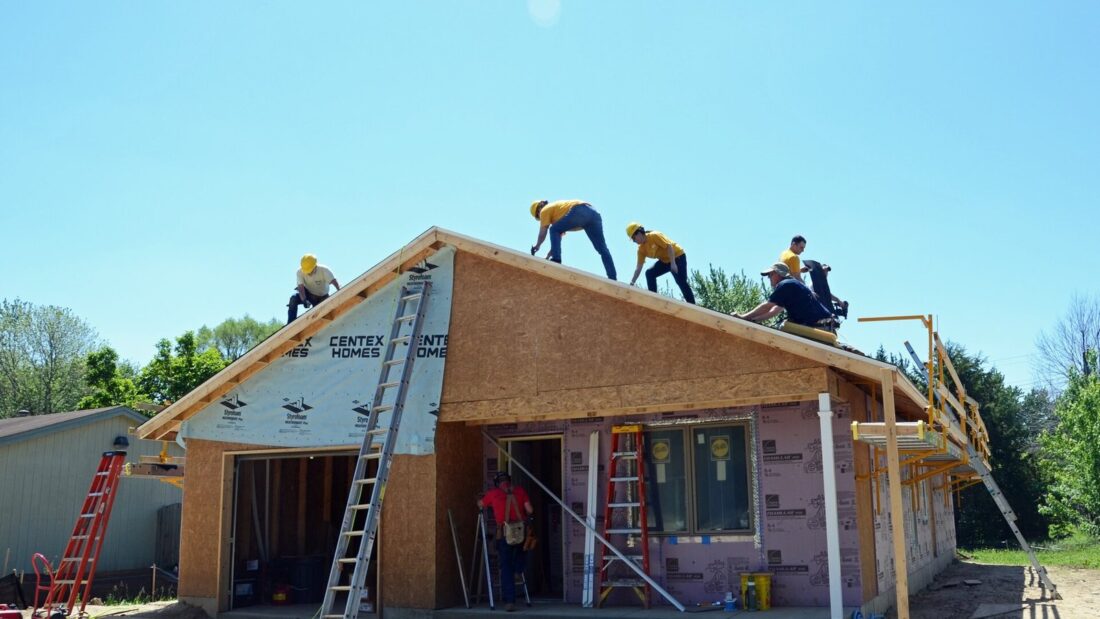 Casa de madeira em construção, ainda sem revestimento, com voluntários de capacete trabalhando no telhado e escadas apoiadas na parede