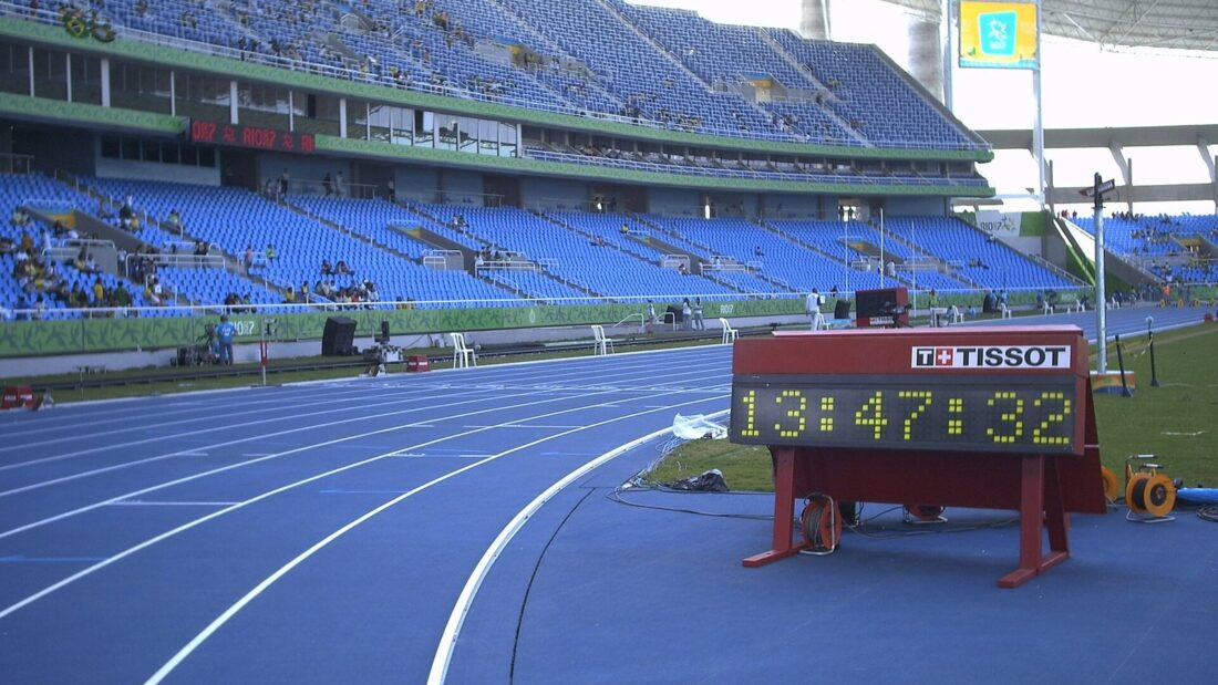 Pista de atletismo azul de um estádio, com arquibancadas azuis e um cronômetro digital vermelho na linha de chegada