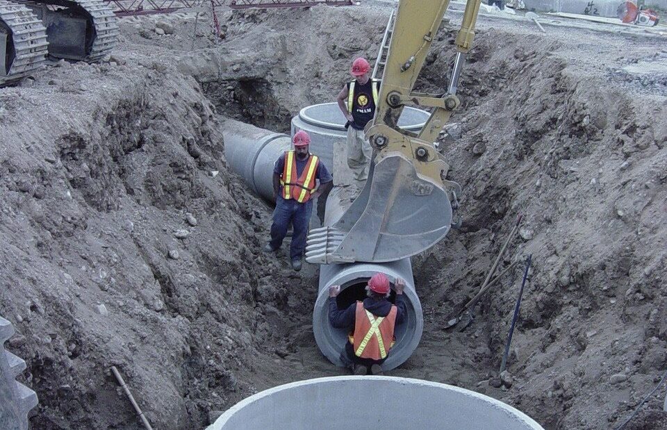 Trabalhadores com colete refletivo dentro de uma vala aberta por escavadeira instalam tubos e poços de concreto de uma rede de galerias de água da chuva