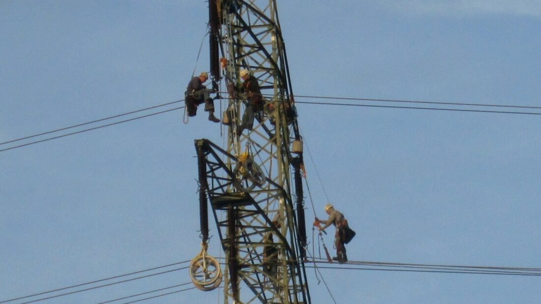 Dois trabalhadores no alto de uma torre metálica fazem manutenção em cabos de uma linha de transmissão de energia, sob céu azul