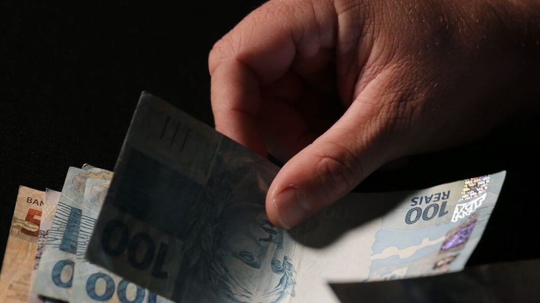 Mão segurando cédulas de 100 reais contra um fundo escuro