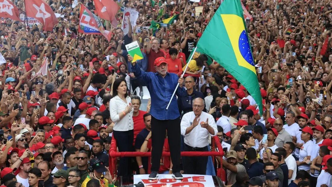 Lula acena de cima de um carro de som, cercado por multidão vestida de vermelho e com uma grande bandeira do Brasil