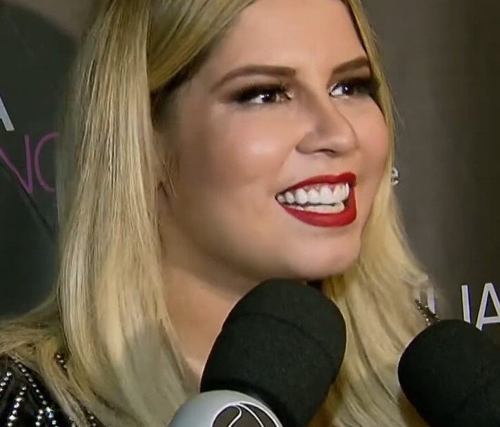 Marília Mendonça sorrindo, de cabelo loiro, diante de microfones de emissoras de TV durante entrevista