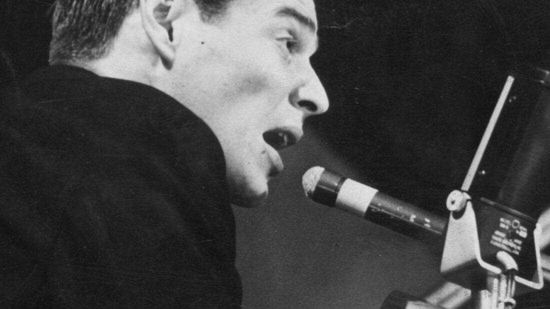Foto em preto e branco de Tom Jobim jovem, de perfil, cantando diante de um microfone