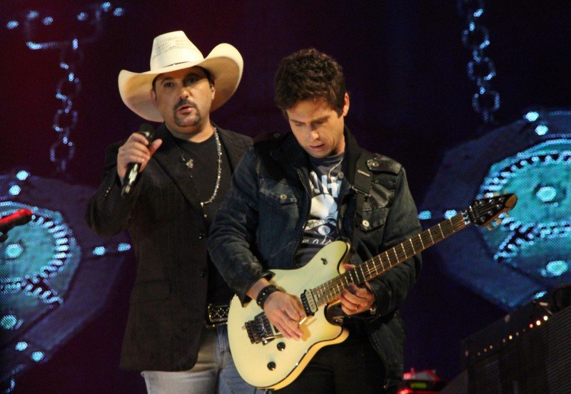 Edson, de chapéu branco e microfone na mão, e Hudson, tocando guitarra, em um palco com luzes azuis