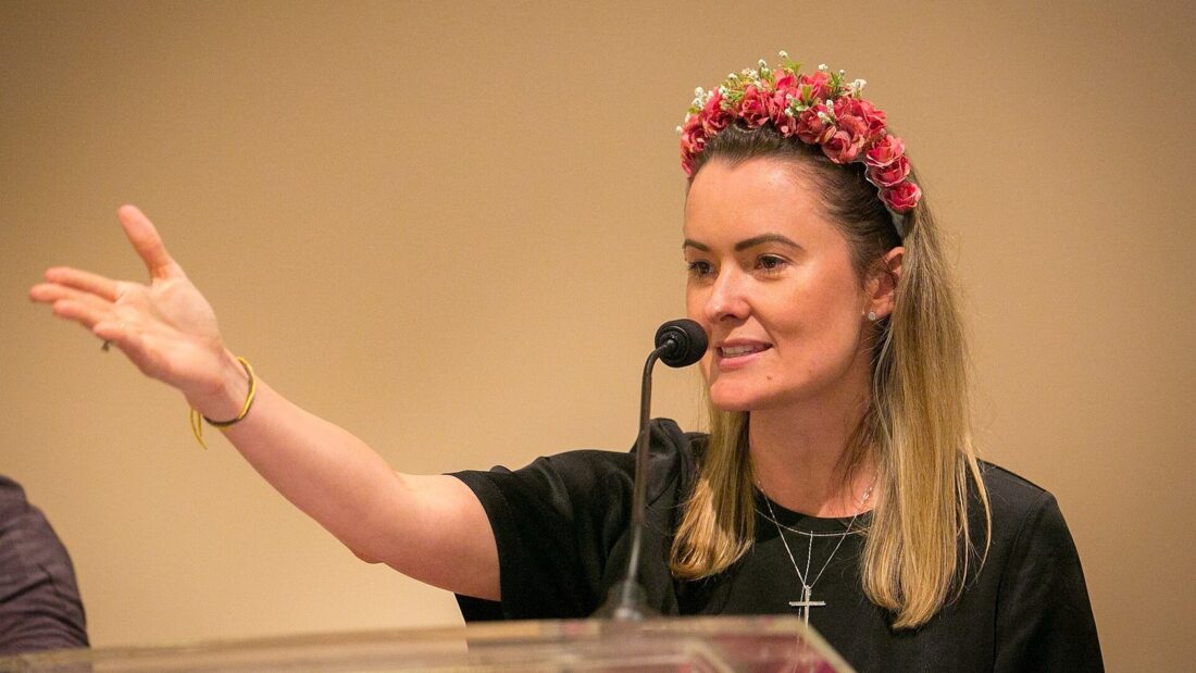 Mulher de cabelos loiros, com uma tiara de flores, fala ao microfone atrás de um púlpito transparente e aponta com o braço estendido