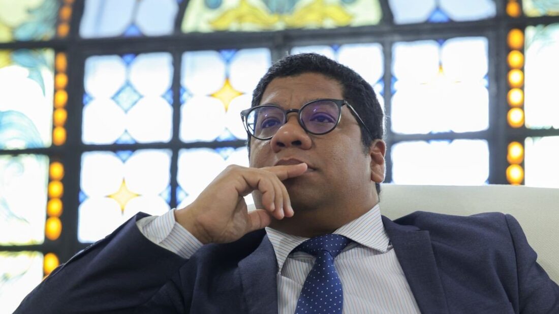 O embaixador de Cuba no Brasil, Victor Manuel Cairo Palomo, de terno e gravata azul, sentado, com a mão no queixo, diante de um vitral colorido