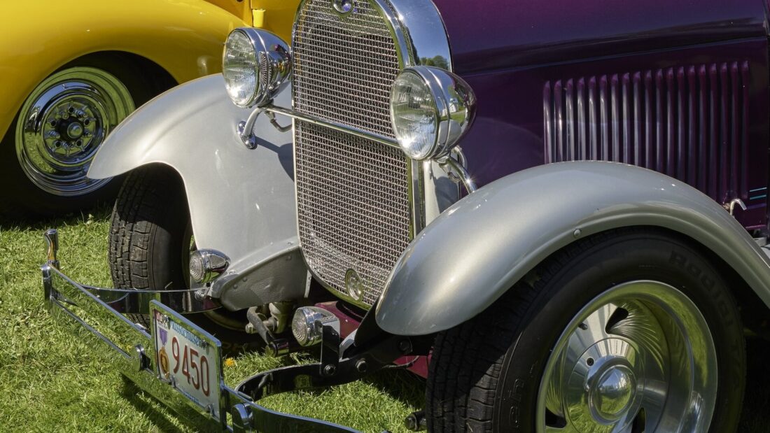 Carro antigo de capô alto, para-lamas cromados e faróis redondos estacionado na grama ao lado de outros carros clássicos em uma exposição
