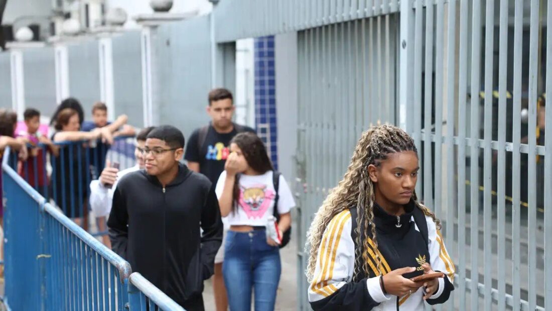 Estudantes, alguns de mochila e com celular na mão, saem pelo portão de grade de uma escola depois da prova do Enem