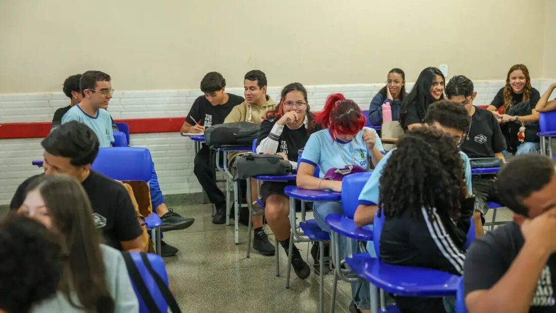 Estudantes do ensino médio sentados em carteiras azuis, em sala de aula, conversam e fazem anotações