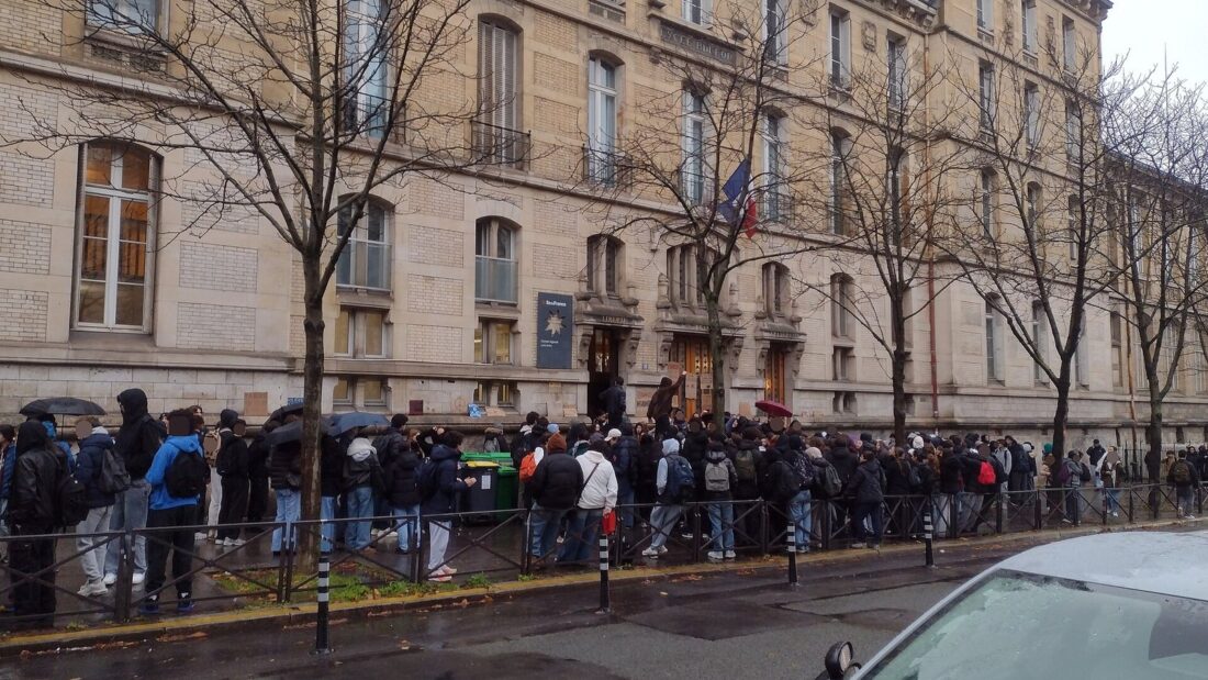 Fila de estudantes diante de um prédio escolar antigo de vários andares em Paris, em dia chuvoso