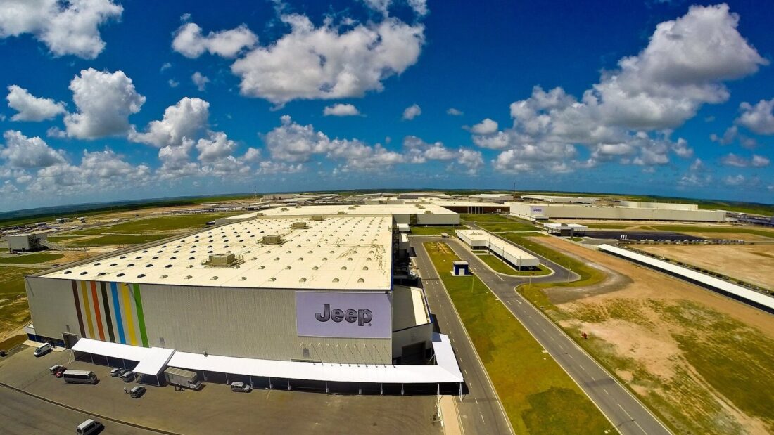 Vista aérea de um grande complexo industrial, com galpões, pátios e vias internas, sob céu azul com nuvens