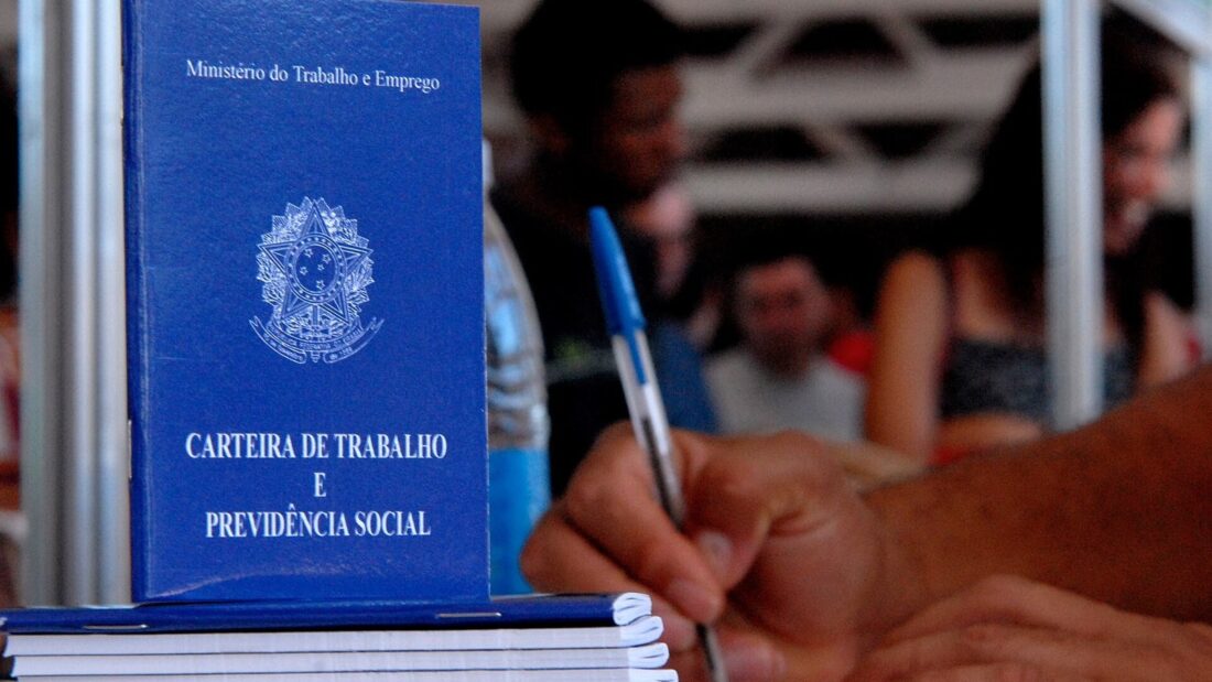 Carteiras de Trabalho e Previdência Social de capa azul empilhadas em uma mesa, com uma pessoa preenchendo um formulário ao lado