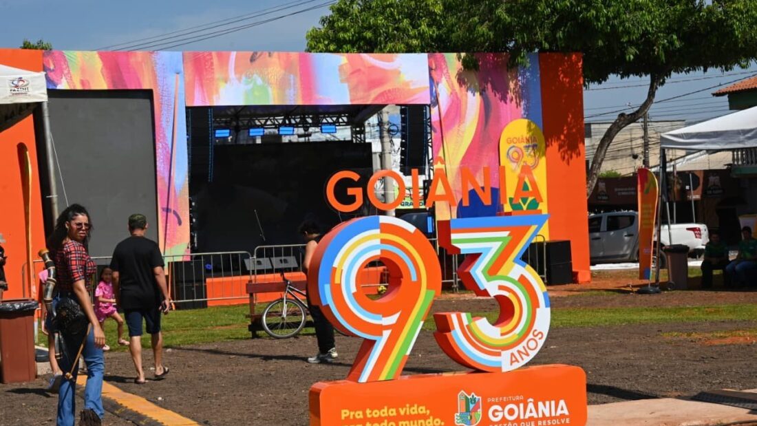 Letreiro colorido com o número 93 e a frase Goiânia, pra toda vida, pra todo mundo, diante de um palco montado com painel de cores vivas, com pessoas circulando