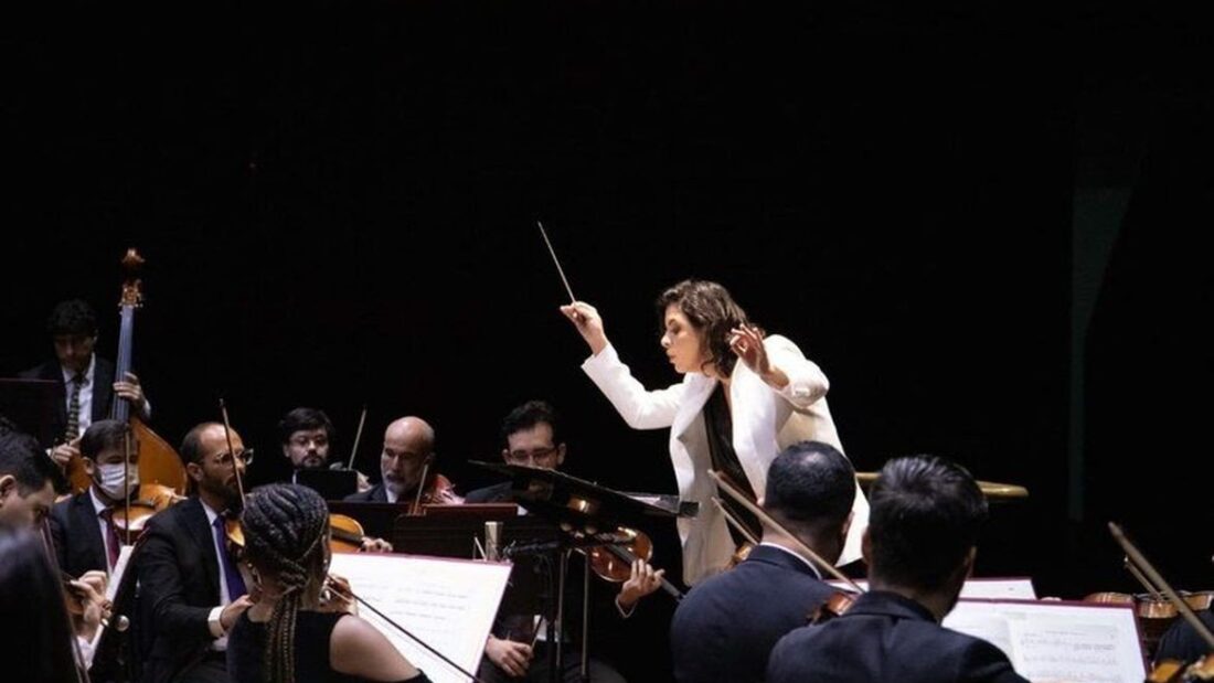 Regente de terno branco conduz músicos de orquestra vestidos de preto em um palco escuro, com violoncelos e contrabaixo à esquerda